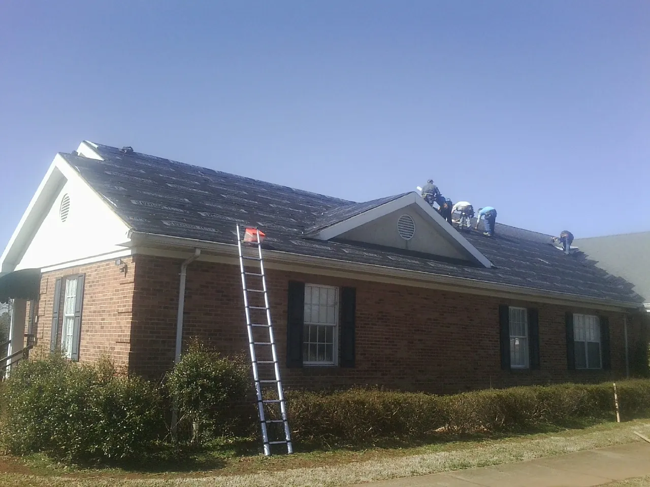 Crew tearing shingles off a brick ranch roof with a ladder set — Emergency Roof Repair work by our crew in New Era