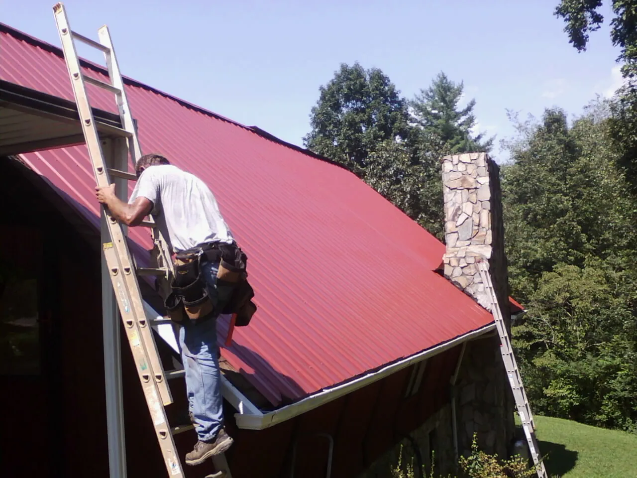 Roofer on a ladder beside a red metal roof and stone chimney — Soffit & Fascia Repair by Roofing Brothers in New Era